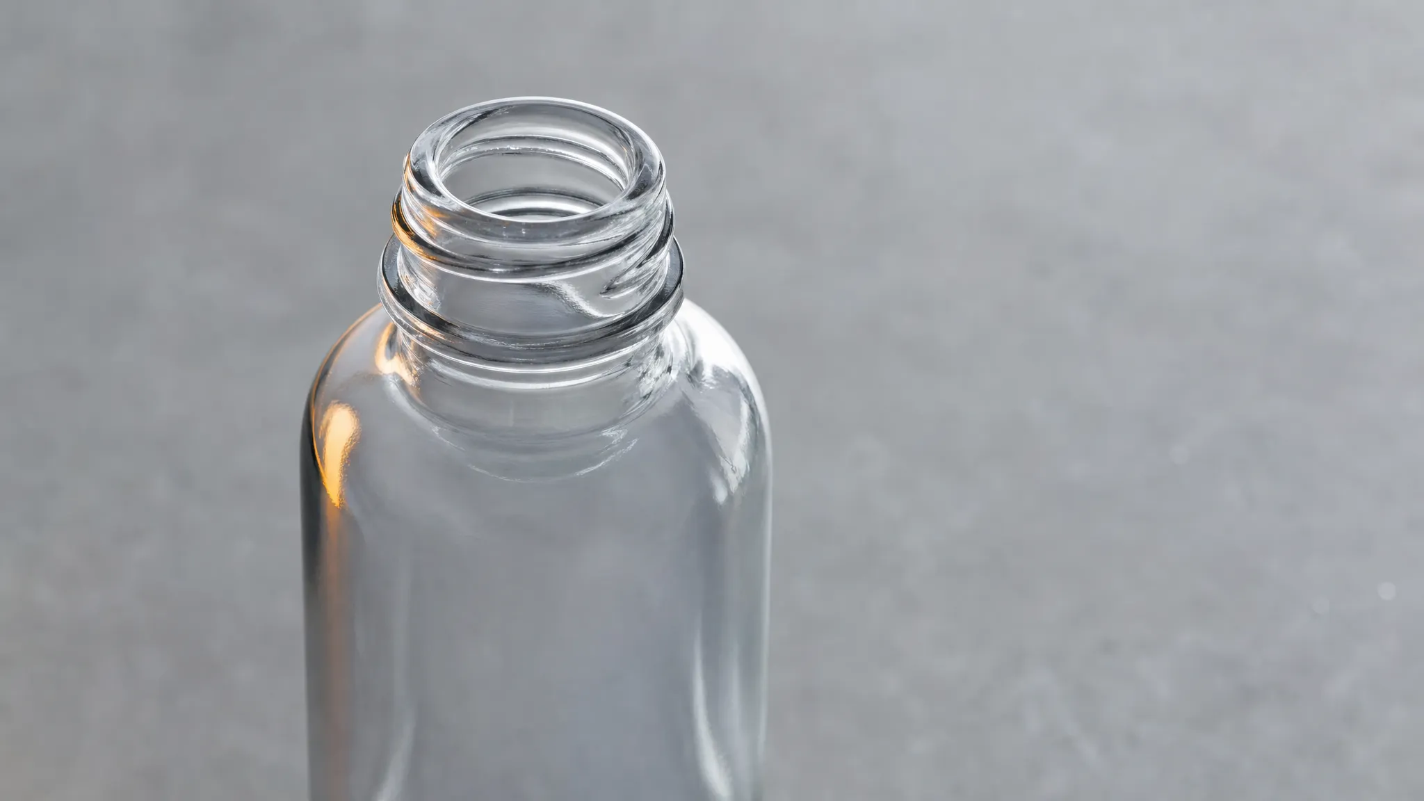 The open neck and mouth of an empty clear glass bottle on a neutral bench, the opening a pour has to reach