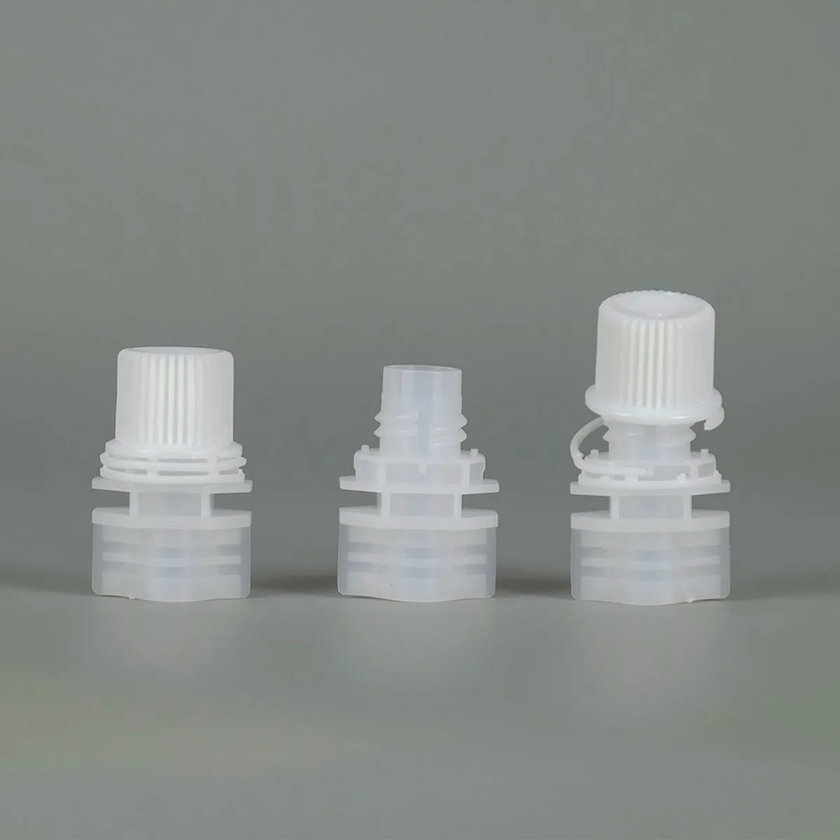 Three white plastic spout caps for stand-up pouches on a grey background, the right-hand sample joined to its spout base by a tether strap