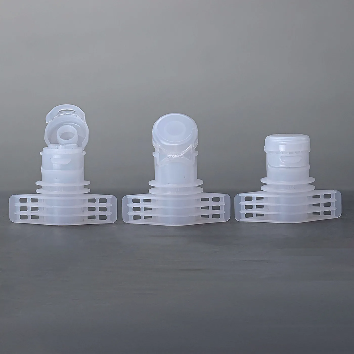 Three flip-top spout caps on a grey surface, the left one open on its hinge and two closed units behind it