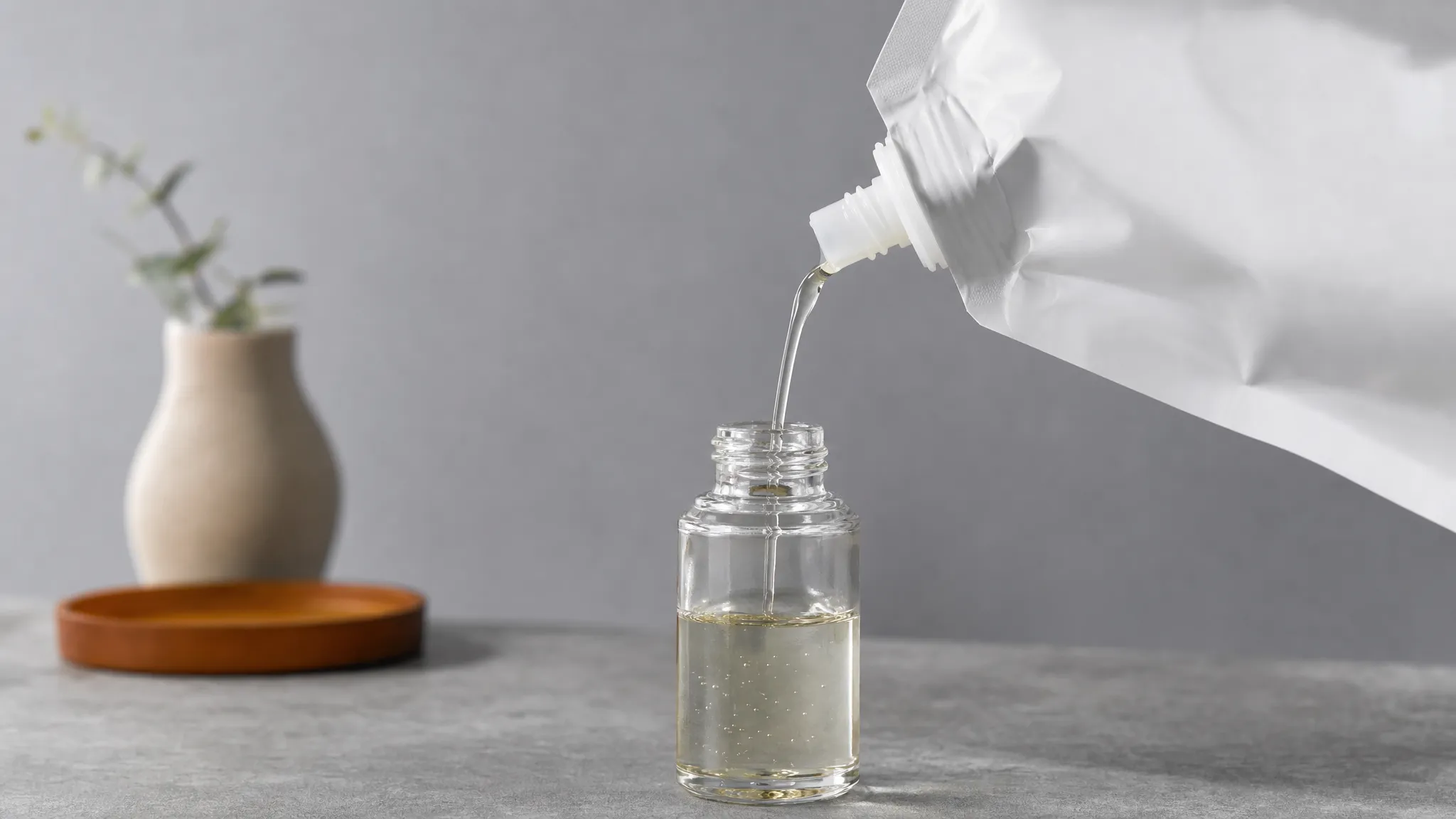 A spouted refill pouch tilted over a small bottle opening, pouring a thin steady stream of liquid on a neutral bench