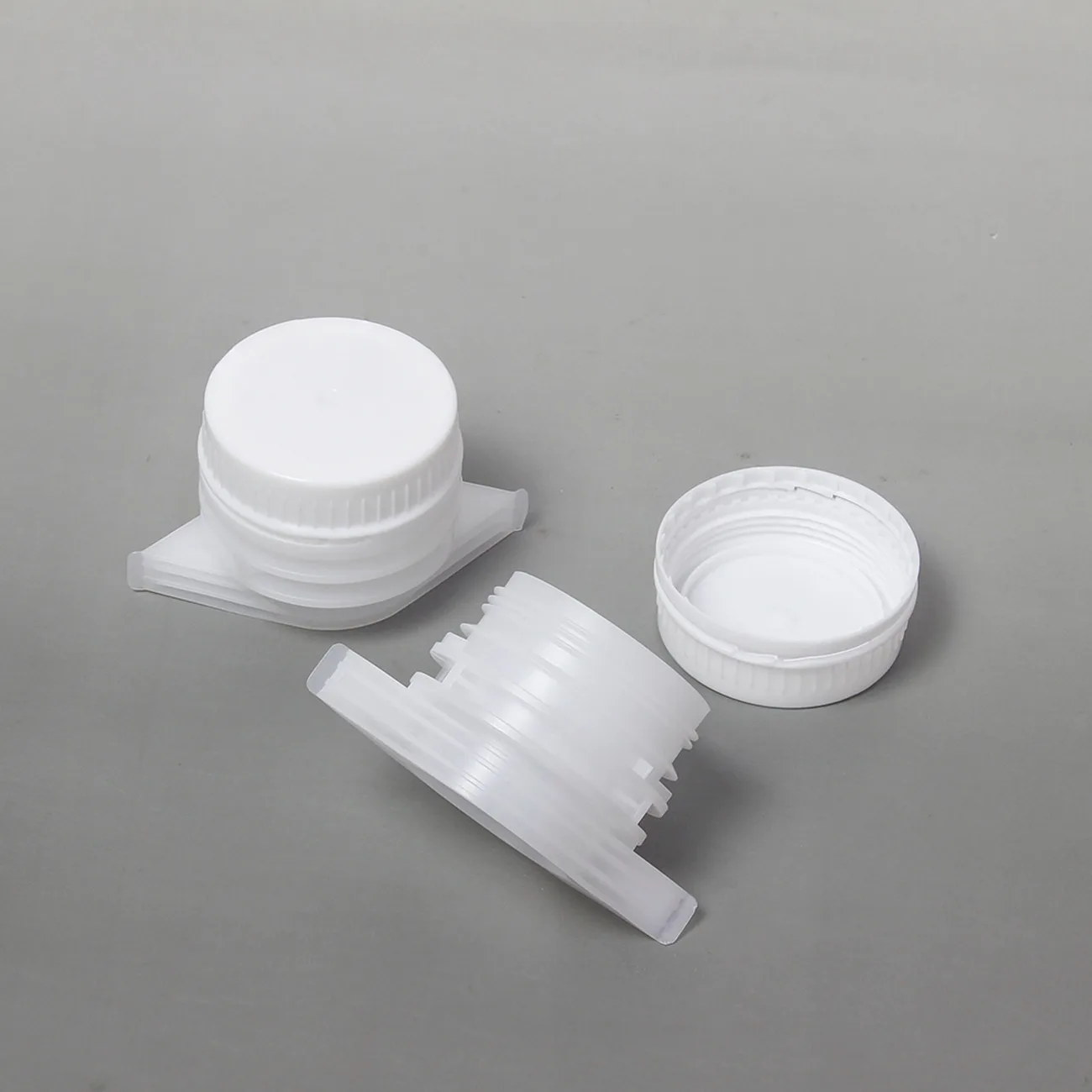 A white plastic screw spout cap with the lid taken off and the bore visible, beside a translucent spout body on a grey bench