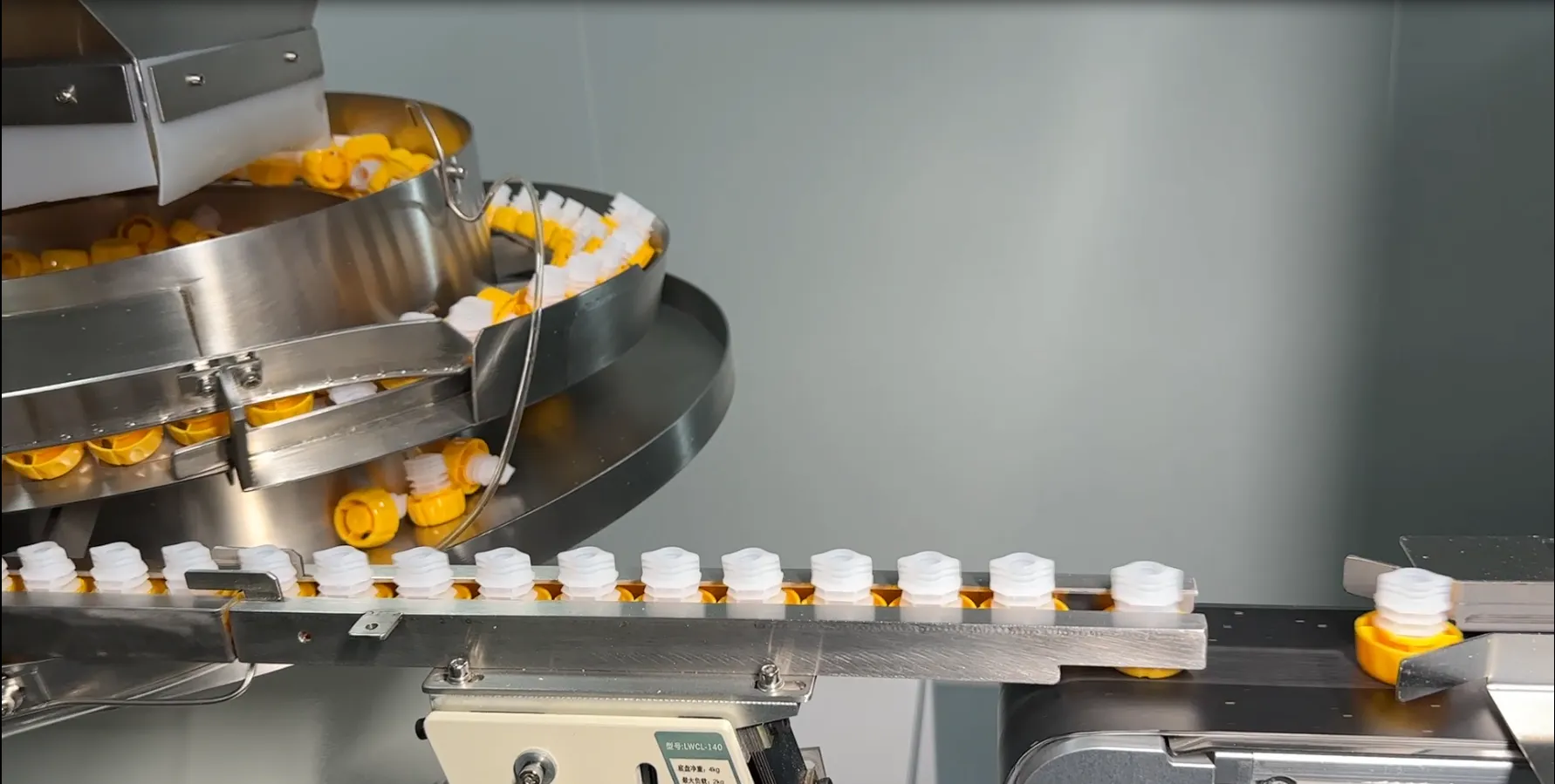 Yellow and white plastic spout caps being oriented in a stainless-steel bowl feeder and fed onto a rail on a packaging line