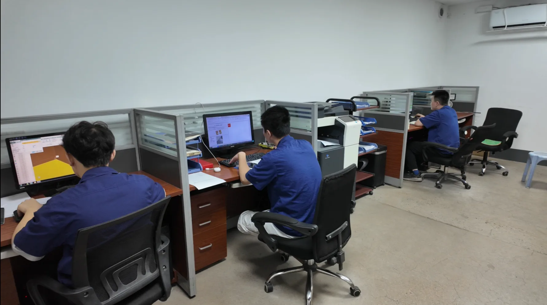 Three people in blue work shirts at computer workstations in an office, one monitor showing a 3D model view