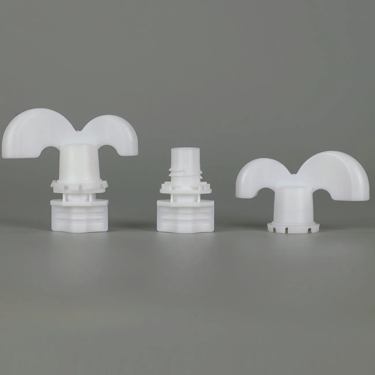 Three white plastic spout caps with wide wing-shaped grips, arranged on a neutral grey background to show the surfaces a hand turns